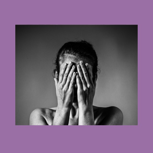 Image of a person pulling up a black sweater over their face in fear. The background is a pale purple.