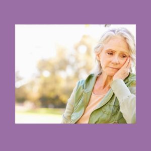 Image of a a person with their hand on their face, with a grief struck look on their face. They have pale skin, white hair, and wear a blue top. The background is a pale purple.