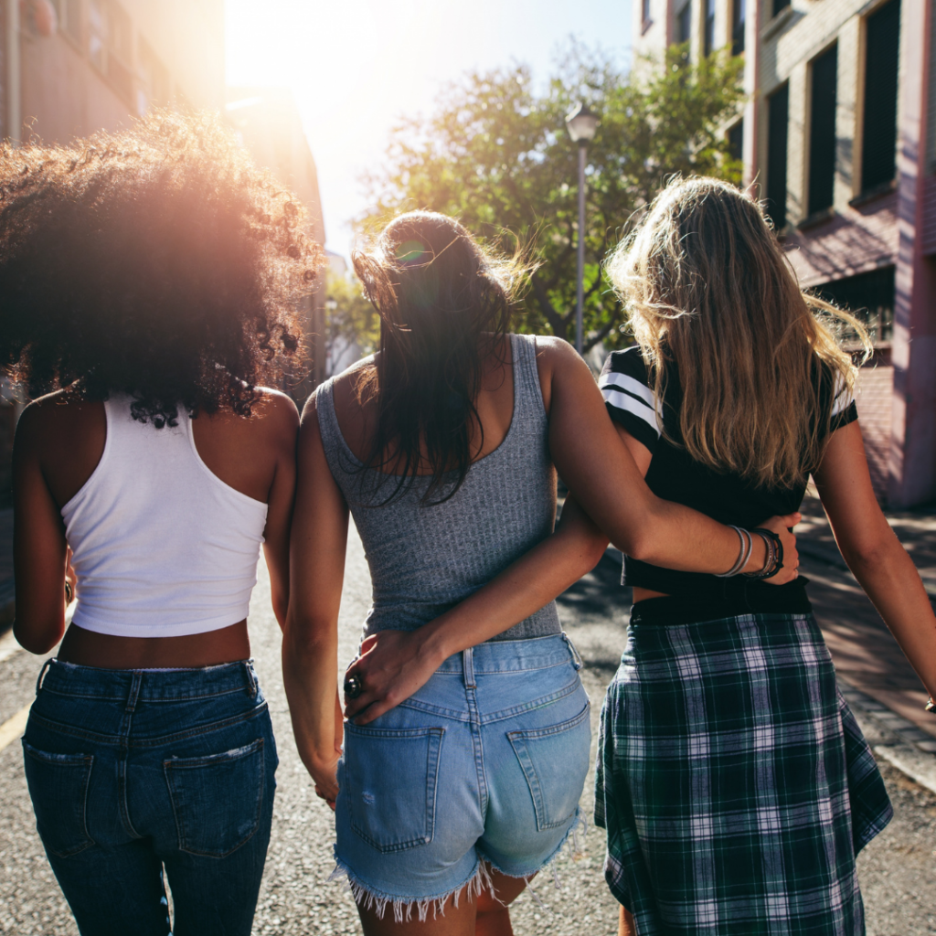 Image of three people from behind, walking down a street with apartments and trees, and the sun radiating down the street. The person on the left has dark hair and skin, a white top, and dark blue jeans. The person in the middle has brown hair and brown skin and is wearing a grey tank top and light blue jeans. The person on the left has blondish hair, white skin, and a flannel skirt. The three people have their arms around each other representing the importance of community when you have Lichen Sclerosus.