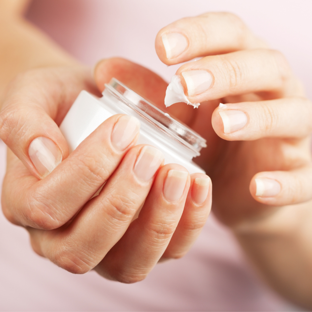 Close up of light skinned hands holding a cosmetic jar of thick cream/emollient. One hand is cradling the jar while the other hand is gingerly dipping a finger or two into the white substance, a small portion of which sticks to the finger tips. The fuzzy background is of the person's pale pink shirt.