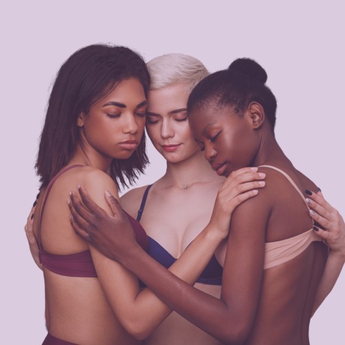 Three people from the waist up wearing only bras embrace. Their eyes are closed and faces are downcast. The first person has shoulder length straight brown hair and light brown skin and wears a burgundy bra. The second person has a platinum blond pixie cut and white skin and wears a navy blue bra. The third person has straight black hair pulled into a high bun and dark brown skin and wears a blush-pink bra. The image represents vulnerability and supporting others.