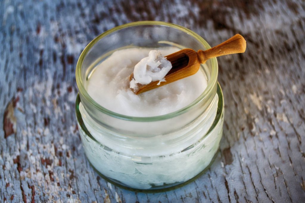 A close up of a short glass jar of solid coconut oil with a small wooden scoop resting on top. The scoop has been used scrape a small portion of oil onto the scoop. The jar rests atop a weathered wooden table with pealing white paint. Coconut oil is my emollient of choice.