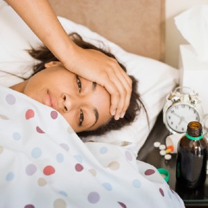Person with brown skin and short black hair lies in bed under a white blanket with colorful polka dots with their hand on their forehead and drooping eyes. On the table next to them are some pills, a bottle of cough syrup, a glass of water, and a tissue box. This represents my taking a little break from dilators when I got sick.