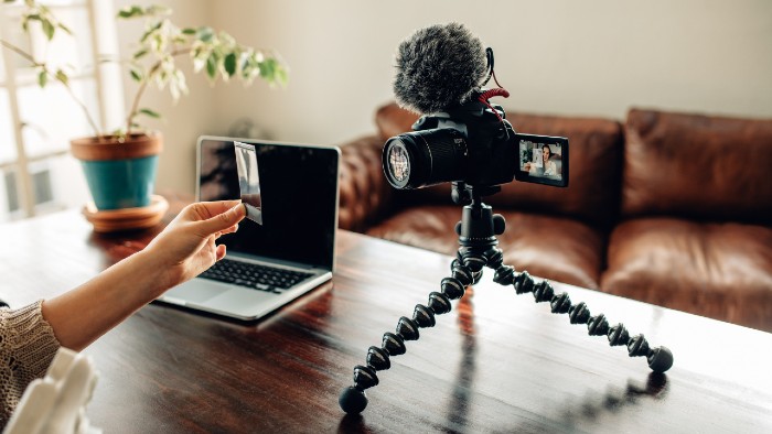 Image of a desk with a recording camera, tripod, mic, and computer representing a YouTube filming set up. There are plants on the desk and a brown couch in the background.