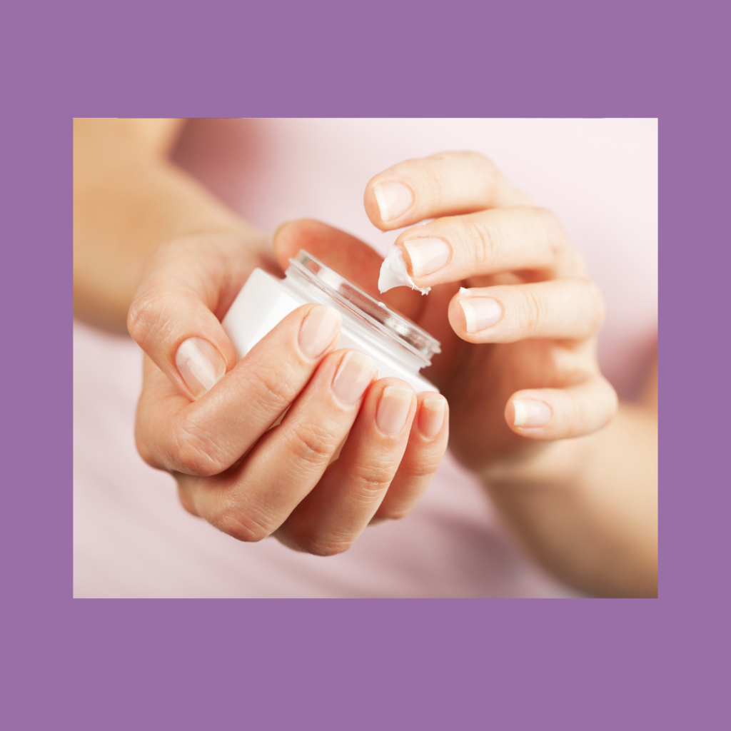 Close up image of light skinned hands with white, short acrylic finger nails holding a cosmetic jar of pink cream/emollient. One hand is cradling the jar while the other hand is gingerly dipping a finger or two into the cream, which now has a short peak from where the fingers withdrew. The fuzzy background shows a person's torso wearing a white robe. There is a light blue frame around the image and a black title at the bottom that reads, "Emollient: One of the Most Overlooked Products for LS".