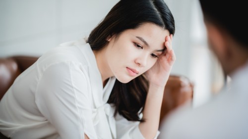 Seated person with white skin and black medium-length hair leaning forward, resting one elbow on their knee and their hand cradling their head around their temple and cheek. Eyes are downcast with a neutral but deep in thought facial expression. The image represents the worry and anxiety that can come from thoughts about perceived health.