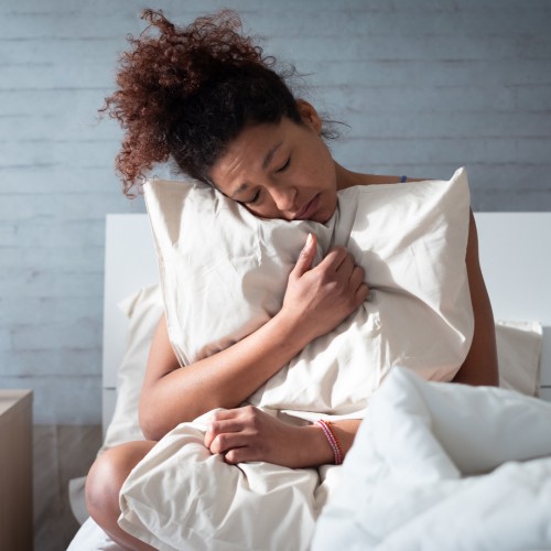 Image of a person with brown skin and brown curly hair in a high ponytail sits up in bed, clutching a white pillow and sheets firmly against their chest with a pained, downcast expression on their face.