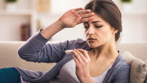 A person with straight brown hair in a low ponytail with olive skin gently places their back hand on their forehead and reclines on a couch, reading a thermometer. Some folks with health anxiety can perceive small changes in bodily sensations, such as body temperature.