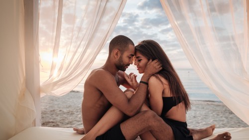 A couple embraces before an open window with loosely drawn curtains that reveal the ocean beyond. They are seated facing each other with knees at 90 degree angles and interlaced with one another. The person on the left has brown skin, a buzzcut, and a short black beard and holds their palm gently against the right side of the other person's jaw line. The person on the right has long straight brown hair and light brown skin and is wearing a black bralette and black shorts. Their foreheads are touching lightly and they both have their eyes closed with slight smiles.
