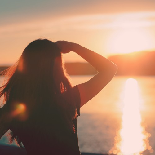Image of a person from behind, with their harm raised and hand on their forehead looking out at a sunset on a beautiful lake.