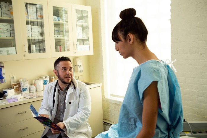 Image of a person with dark hair in a bun in a hospital gown sitting on a table while doctor explains her diagnosis to her.