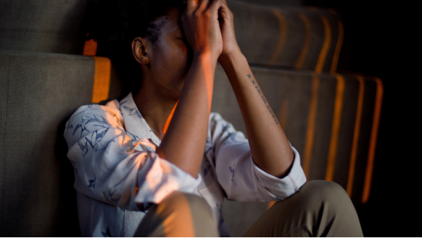 Image of a person with dark skin and short dark hair, sitting on a floor with their hands up over their face expressing grief and loss.