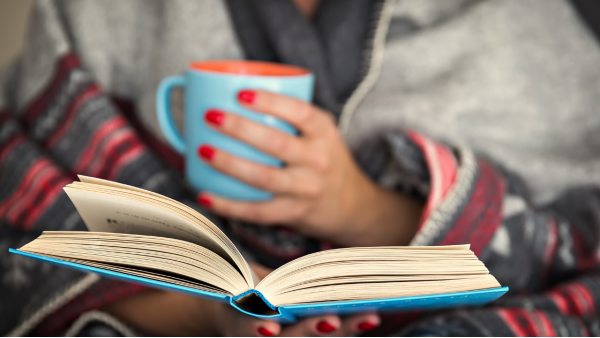 Image of a person in a grey sweater, holding a teal mug with bright red-coral nails reading a book.