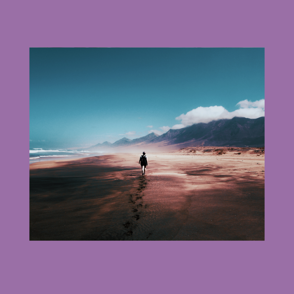 Person walking into the distance down a very long beach. There are great mountains to the right with several fluffy white clouds at the peaks. To the left, gentle waves are breaking against the shore. A medium thickness purple border surrounds the image. This represents my dilator journey.