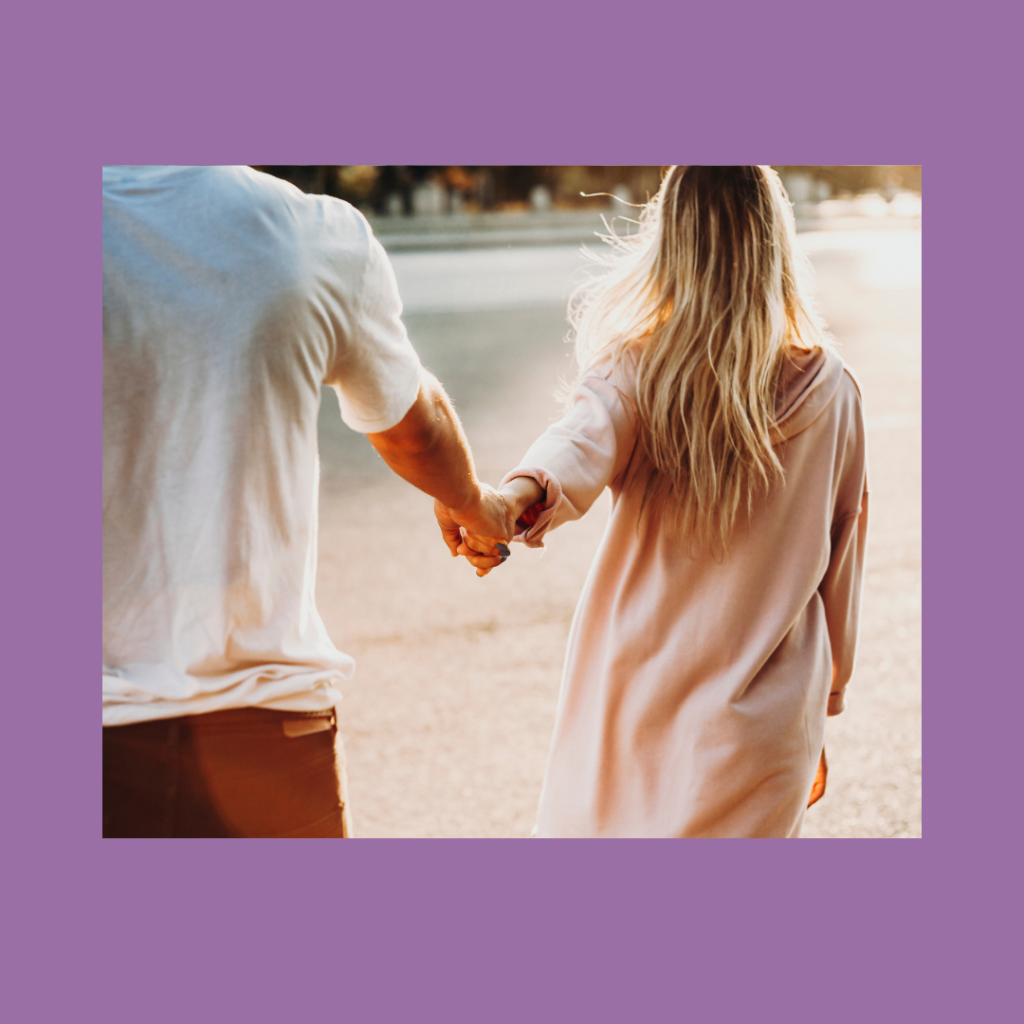 Backside view of two people walking together across a field. The person with long bleached blond hair with brown roots and an oversized blush colored sweater dress is leading by hand the person with a white short sleeved t shirt and dark denim jeans.  The image is surrounded by a soft medium-thickness purple border