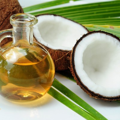 Golden oil in a round glass jar with a narrow spout lays beside a split open coconut atop a coconut leaf on a white table.