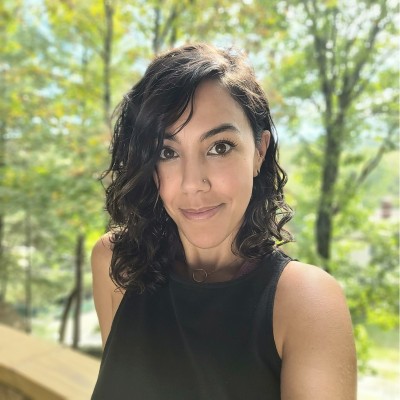 Image of Dr. Yaghmail wearing a black halter top with wavy brown hair to her collarbone. She is standing outdoors with trees in the background. 