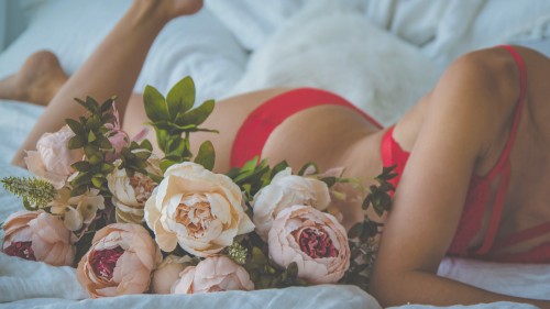 Image of a person lying on their stomach in a white bed wearing a red lacy bra and matching underwear with a bouquet of pale pink flowers lying beside them. This image represents the intimate nature of this post about my dilator journey.