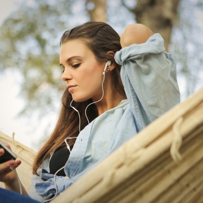 A person with long brown hair and light skin relaxing in a swinging hammock. They are wearing a black tshirt and an oversized denim button-up with the sleeves rolled up to the elbows. They are holding their smartphone with one hand and rest their other hand on the back of their neck with elbow sticking out. Their eyes are downcast to their phone and they have a calm, relaxed facial expression as they listen to music using white earbuds. Listening to a podcast/music can be a great way to calm nerves before or during a vulvar biopsy.