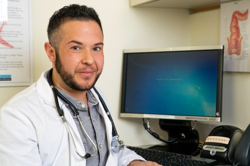 Doctor in a white lab coat with stethoscope around their neck smiling warmly at the camera. They have a medium-length beard and a fade hairstyle. They are seated at a computer work station in an exam room, and on the wall are medical diagrams. This image represents talking to your doctor about your vulvar biopsy. Image taken by Zackary Drucker, The Gender Spectrum Collection.