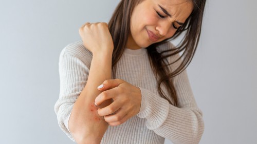Person with long brown hair wearing a white ribbed sweater scratching an itch on their right elbow. They have their eyes closed and head bent with a grimaced facial expression as they scratch the itch hard. The image represents how itch is a lichen sclerosus symptom.