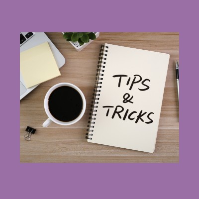 A notepad on a wooden desk with 'tips and tricks' written with a bold, black marker. Scattered beside the book is a cup of black coffee, a pen, a laptop, a stack of sticky notes, and a mini binder clip.