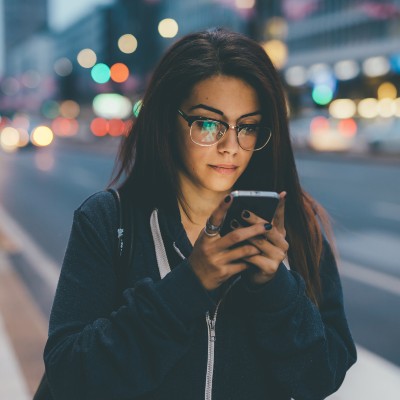 A person with long dark hair and black rimmed glasses walking along a blurry street with the light from their phone on their face.
