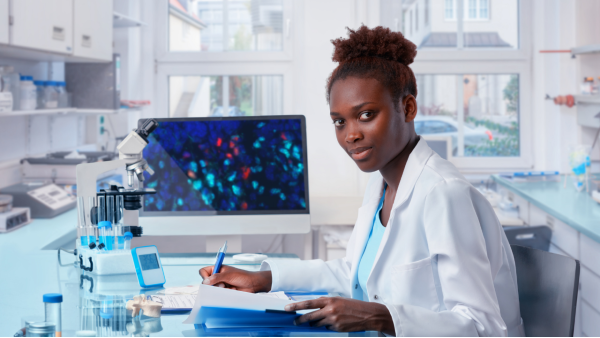 Image of a person with dark brown skin and brown curly hair pulled into a short ponytail wearing a white lab coat working at a pathology desk analyzing biopsy samples.