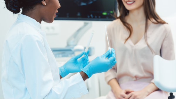 Image of a doctor with dark brown skin and a white lab coat using blue-gloved hands to demonstrate a lab test to a patient with light skin and long brown hair seated and smiling in an exam room. The patient wears a beige v-neck blouse with pale pink slacks.