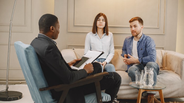 Image of a person with dark brown skin, short black hair in a navy top and pants sitting on a chair with two people, one person with pale skin and short brown bobbed hair in a white sweater on the left and another with pale skin, short brown hair, a white T-shirt and jeans on. This represents two people in a relationship talking to a sex therapist.