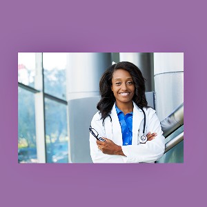 Image of a doctor with brown skin and brown hair in a blue shirt with a white lab coat on standing with their arms crossed, with a smile on their face in a hospital.