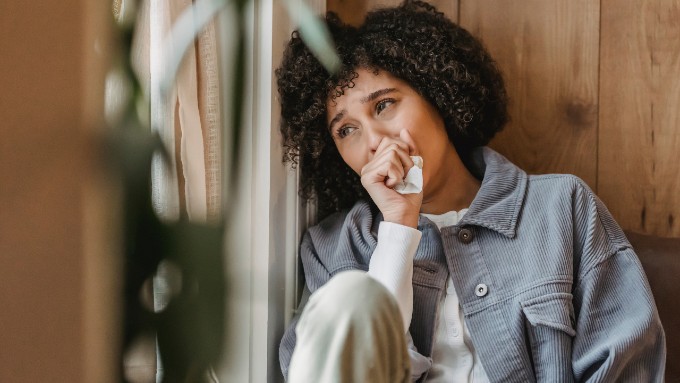 Image of a person with light brown skin and short brown hair sitting with their legs pressed up against their chest. They have a hand holding a kleenex over their mouth as they stifle tears.