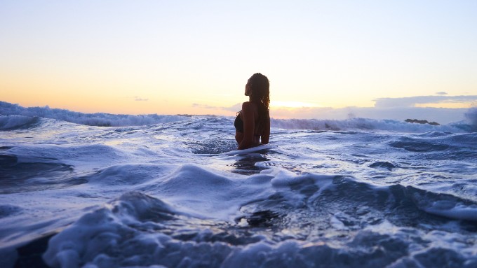 Silhouette of a person with long dark hair from a side profile. They are standing and rejoicing in a beautiful ocean at sunrise. This represents a healing part of Pernille's journey with grief.
