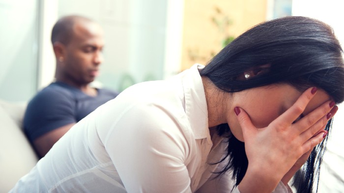 Image of a person with a shaved head, brown skin, and grey-blue t-shirt looking upset and another person sitting next to him with long black hair, tan skin, a white blouse, and their hands over their face as if they are having a difficult and strained conversation.