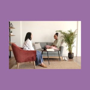 Image of a person with shoulder length brown hair, a white top, and jeans, sitting on a brown chair. Across from this person is another person with brown skin, a grey shirt, sitting on a blue couch with a potted plant next to it. This image represents a patient and a sex therapist discussing sexual health and lichen sclerosus.