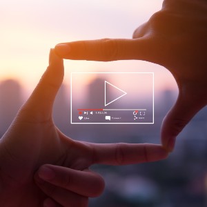 Image of two hands making a square shape with their fingers. Inside the square, which makes it look like a video camera, is a graphic design of a YouTube video